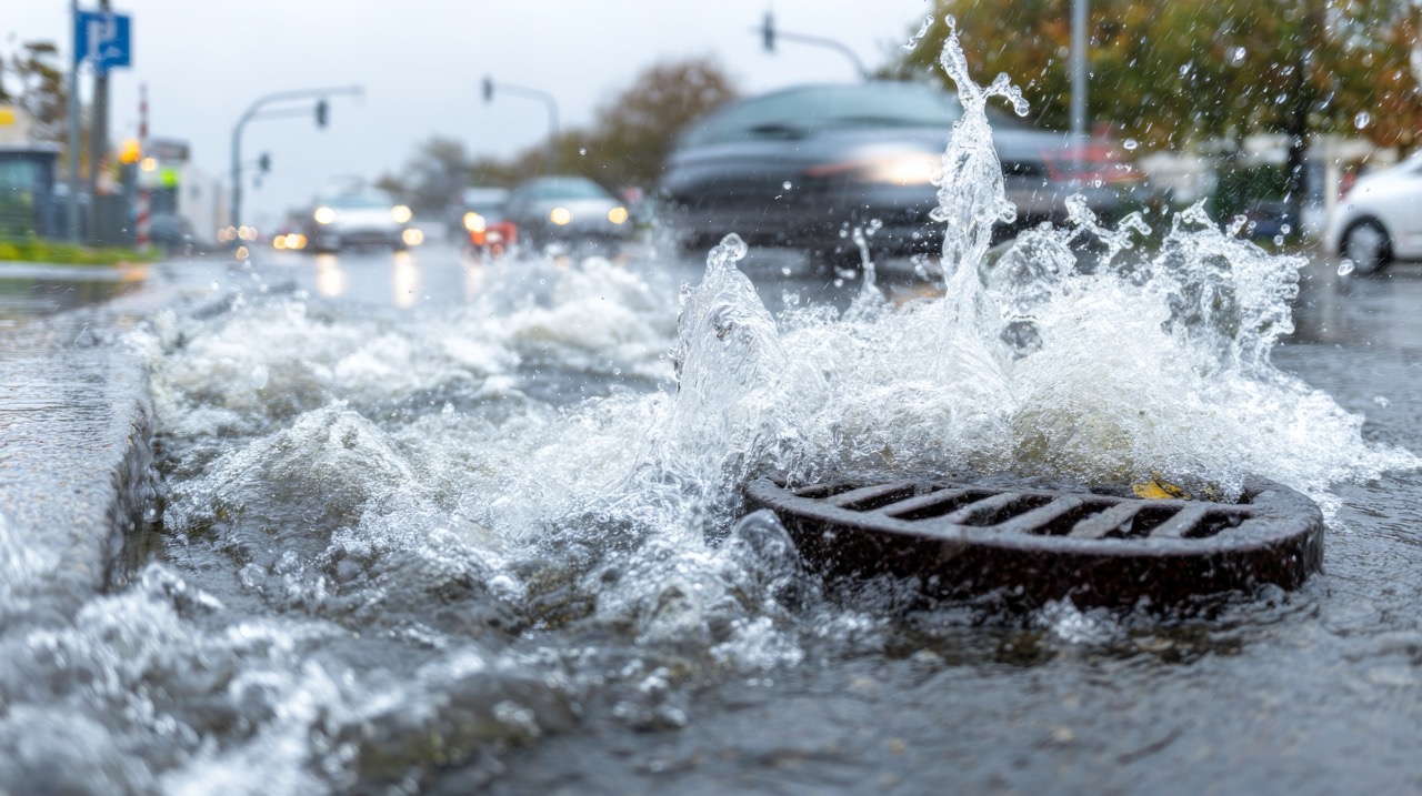 Stormwater rushing down a street and overwhelming a storm drain with cars in the background