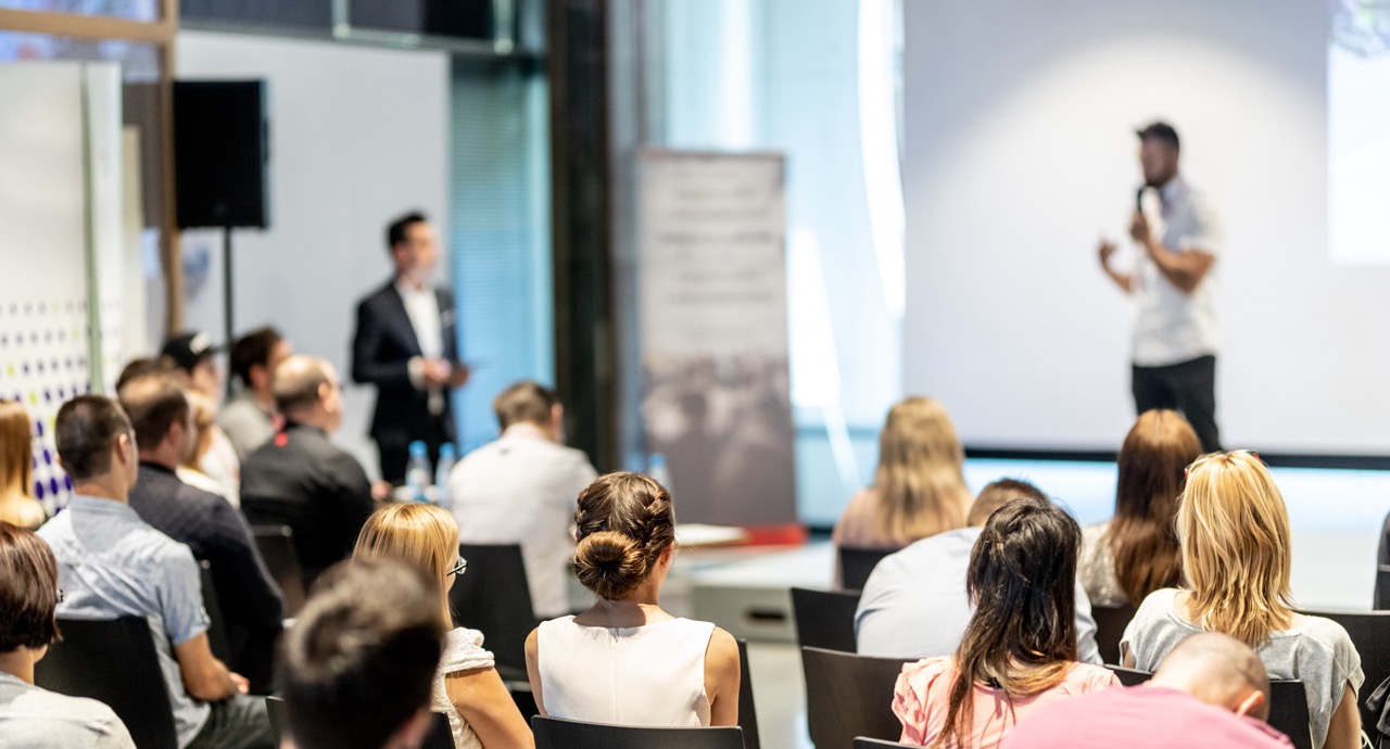Stock photo of a workshop.  Speaker giving a talk at business meeting. Audience in conference hall. Rear view of unrecognized participant in audience.