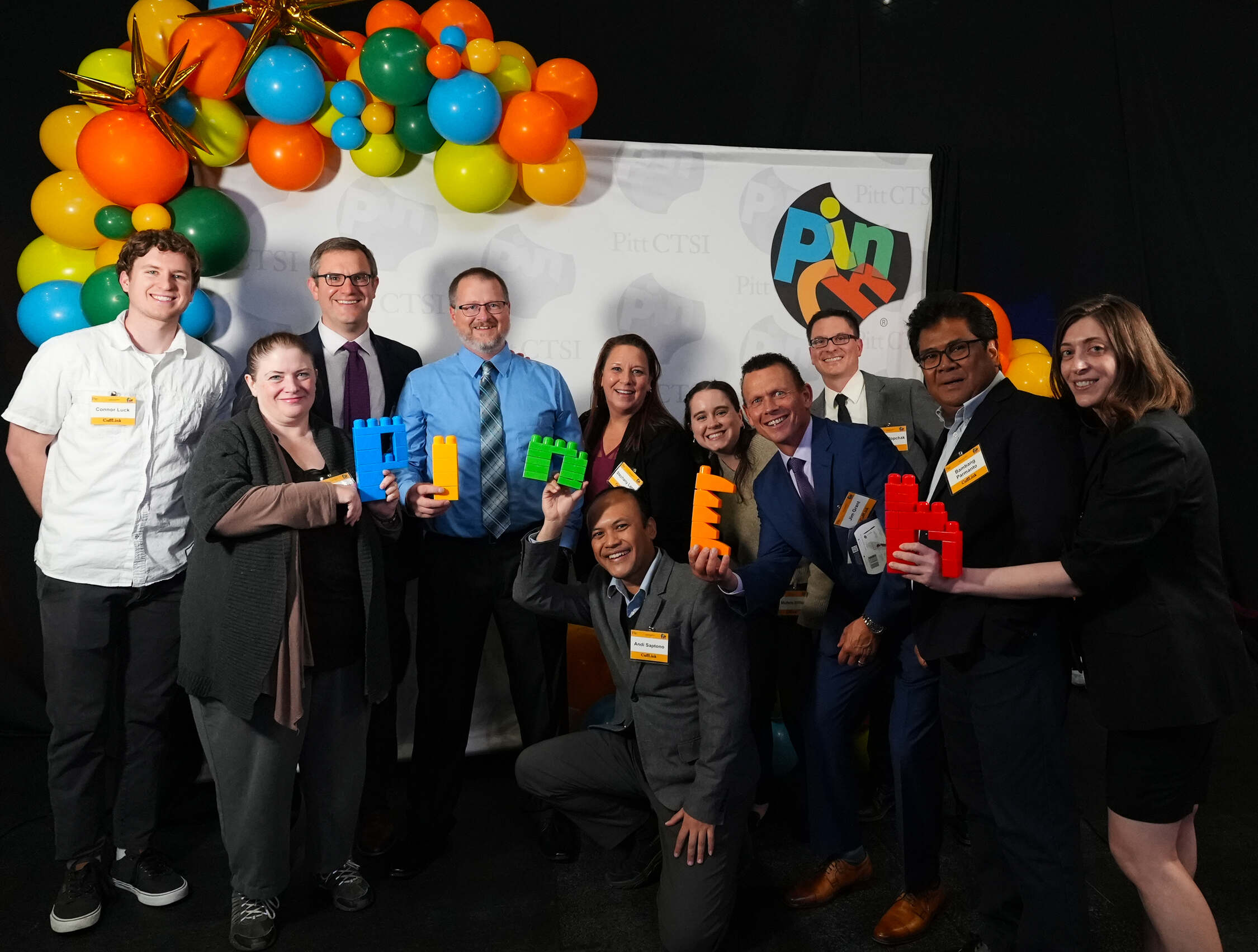 A group of eleven individuals pose in front of a black background, with some holding letters that spell out "PINCH".